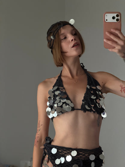 A woman takes a mirror selfie wearing a black halter bikini top and matching bottoms covered in silver coin embellishments, with a matching headband. She holds a pink smartphone to take the photo against a plain gray wall.